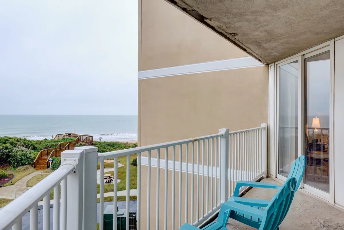 The image showcases the condo's private balcony featuring inviting blue chairs overlooking a breathtaking ocean view. The scene is tranquil, with the vast expanse of the Atlantic stretching to the horizon, perfect for relaxing, reading, or taking in both sunrise and sunset. The mood is calm and serene, suggesting a peaceful retreat steps from the beach.