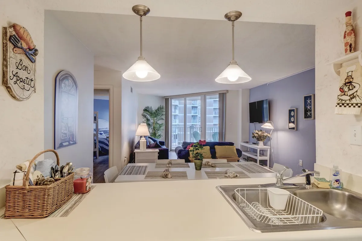 This image offers a comprehensive look at the integrated living, dining, and kitchen spaces. A kitchen island and dining table are visible alongside a sofa, with large windows providing ample natural light. Decor elements reinforce a sense of place and togetherness.
