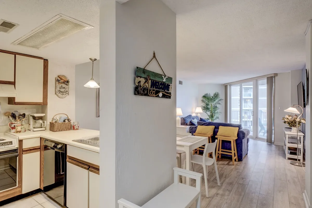 This image presents a well-lit, open-concept interior that seamlessly brings together the kitchen, dining area, and living room. The kitchen area is modern and practical, the dining table is set for a small group, and the living room features inviting seating and coastal-themed decor. A welcome sign and plant add warmth and a sense of arrival, emphasizing the space as the heart of the condo for gatherings and daily life.