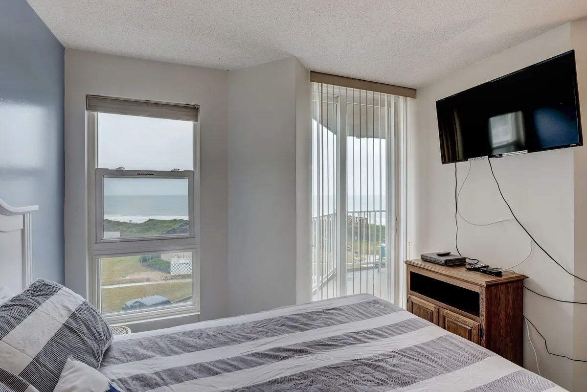 A tranquil bedroom with a striped bedspread is featured, along with a large window offering an expansive view of the ocean. The room is bright, calm, and oriented toward the water, inviting relaxation from sunrise to sunset.