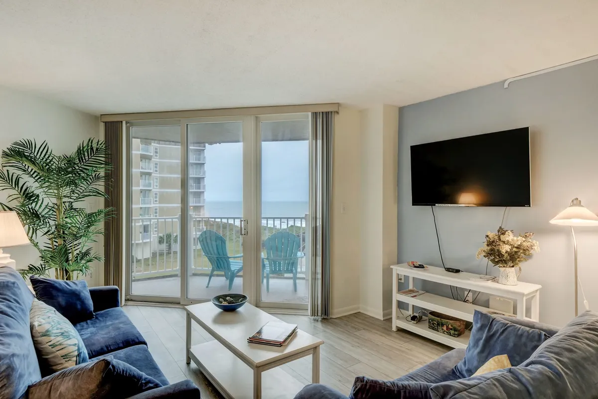 A cozy living area is visible, featuring a sofa, coffee table, television, and sliding glass doors that lead out to the balcony with outdoor seating. Through the doors, the ocean view is just beyond, suggesting seamless indoor/outdoor flow.