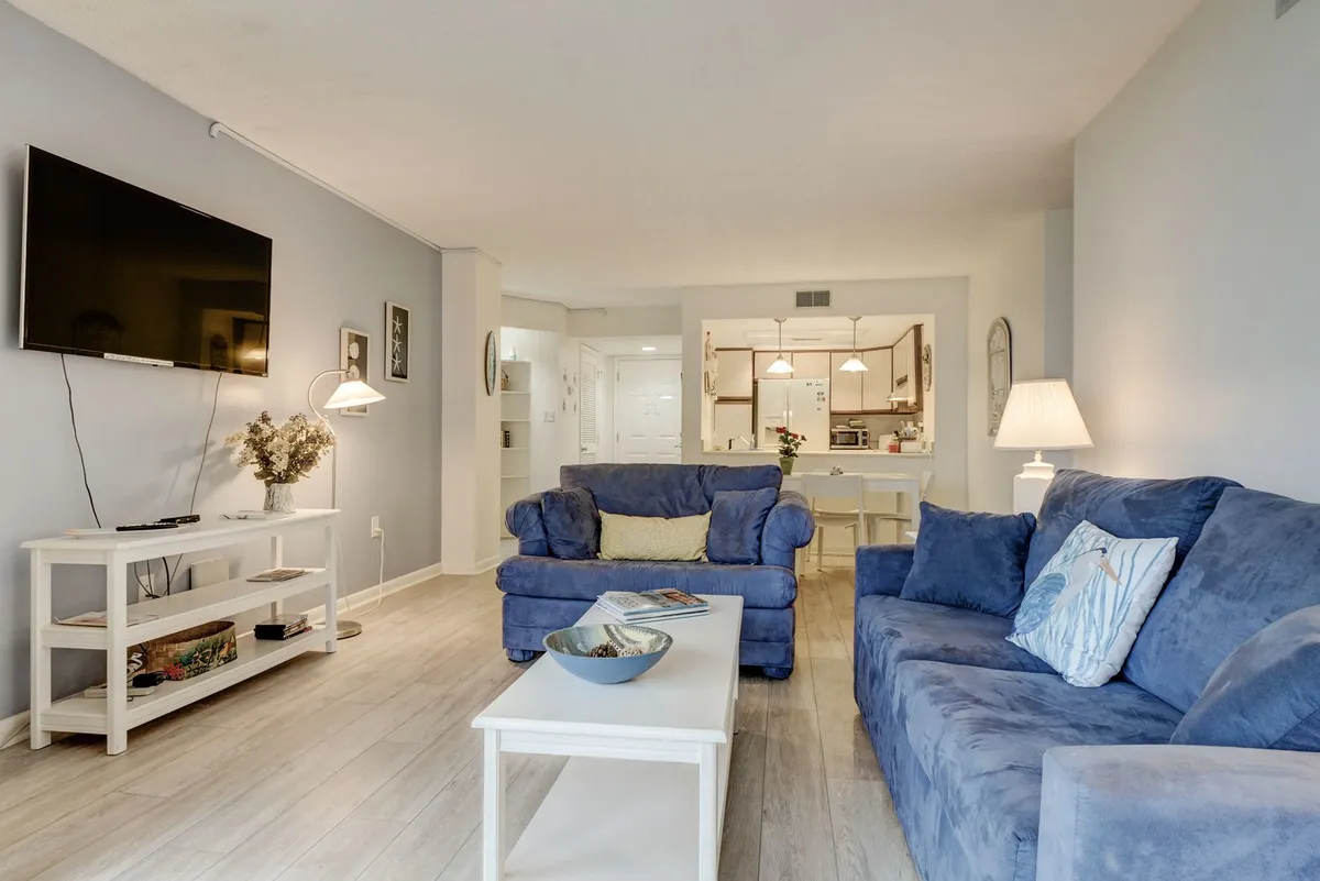 The photo features a cozy living room setup with plush blue sofas, a central white coffee table, and a TV mounted on the wall. Thoughtful lighting and decor create a relaxing and inviting mood, perfect for unwinding or spending time with family and friends.