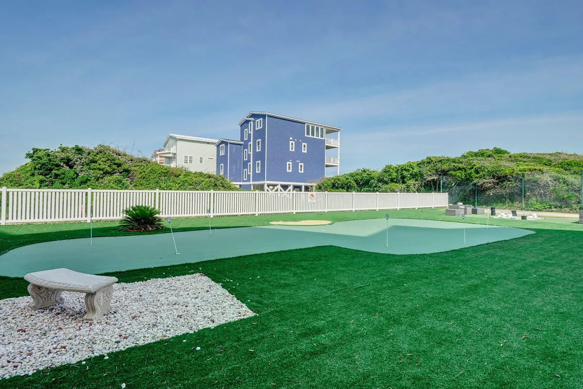 A vibrant outdoor putting green surrounded by lush greenery, a stone bench, and a blue house. The scene is recreational and relaxed, inviting guests to practice golf, socialize, or host casual outdoor get-togethers.