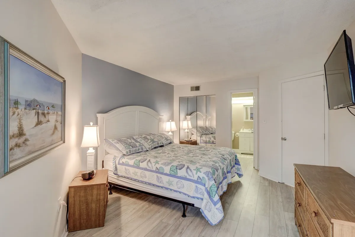 A serene bedroom featuring a bed with patterned bedding, framed artwork above the bed, warm lighting from bedside lamps, and a wall-mounted television. Soft natural light filters into the room, creating a tranquil atmosphere perfect for relaxation after a long day.