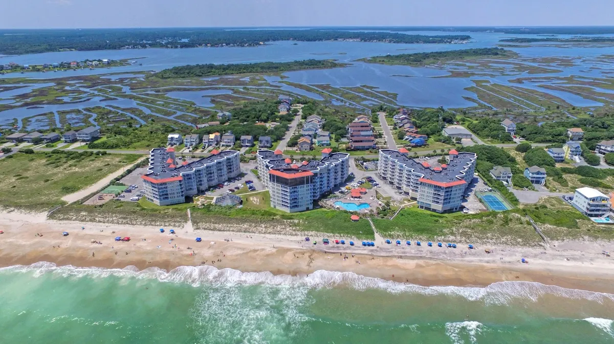 This image showcases a beachfront property with a swimming pool, colorful beach umbrellas, and views of the surrounding marshland. The vibe is vacation-like and carefree, with inviting outdoor gathering spaces perfect for families and friends to relax, play, and enjoy the oceanfront setting.
