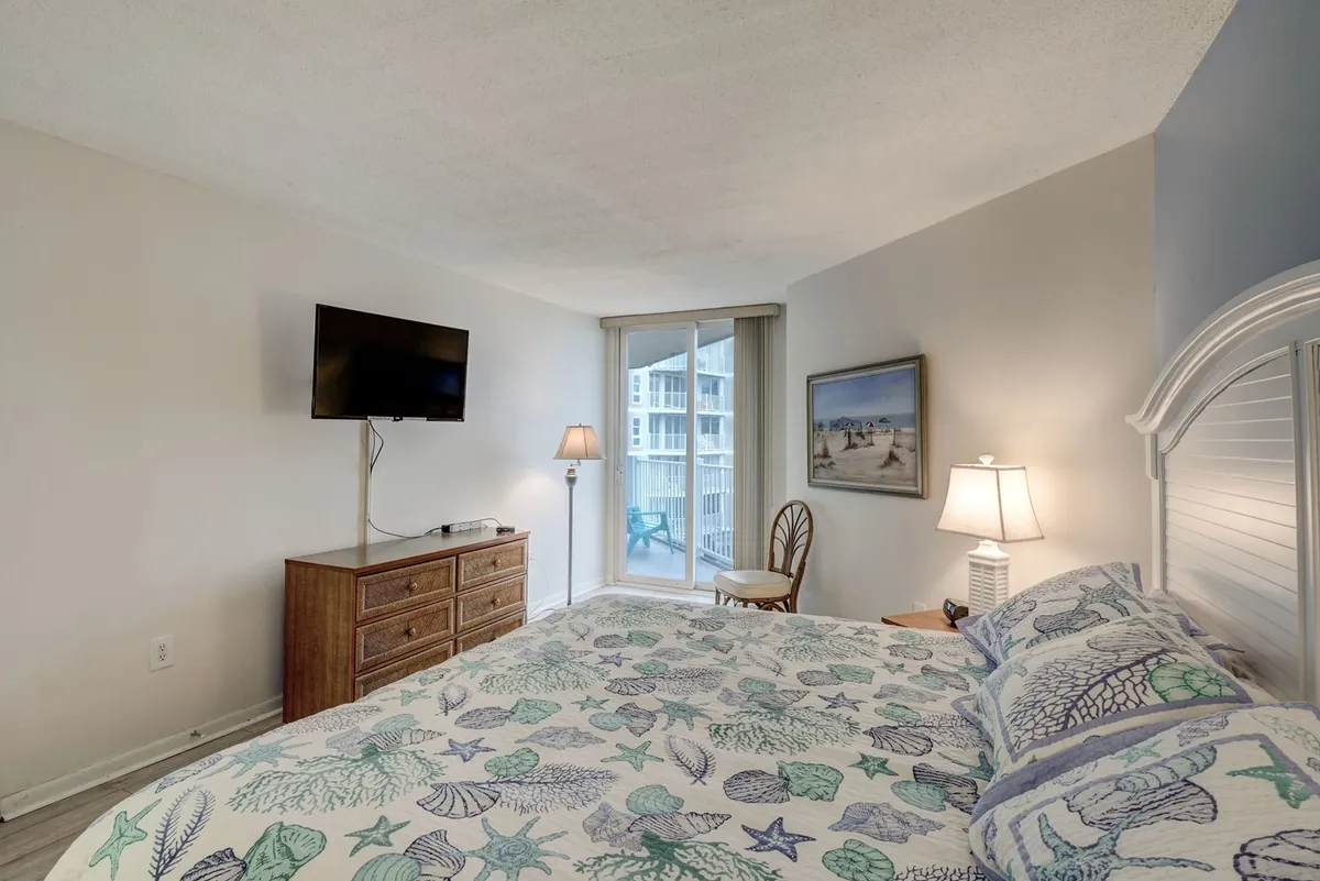 A tranquil bedroom with a sea-themed bedspread, television, dresser, lamp, and glass doors that open to a balcony with ocean views. The space is filled with natural light, offering a peaceful retreat with direct access to the outdoors.